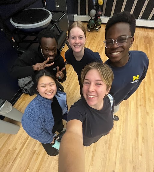 Five students of various ethnicities take a selfie; woman's arm reaches up (group stands on blonde wooden floor).