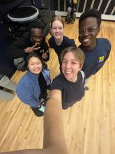Five students of various ethnicities take a selfie (woman's arm reaches up; group is standing on a blonde wooden floor).